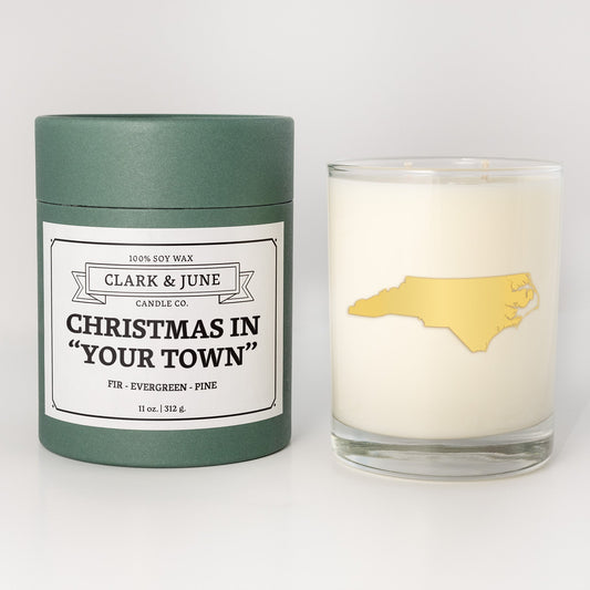 Double wicked soy candle in a 13.5 oz tumbler with the state of   North Carolina printed in 22k gold foil on the face. Green cylinder packaging with Christmas in "Your Town" on the label. SEO Text – Drinking glass, soy wax candle, Christmas Candle, hand poured, small batch, scented candle, Woman Owned, local candle, Housewarming present, gives back, charity, community candle, becomes a cocktail glass, closing gift.