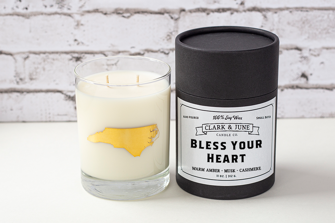 Double wicked soy candle in a 13.5 oz tumbler with the state of North Carolina printed in 22k gold foil on the face. Black cylinder packaging with “Bless Your Heart” on the label. SEO Text – Drinking glass, soy wax candle, North Carolina candle, hand poured, small batch, scented candle, Woman Owned, local candle, Housewarming present, gives back, charity, community candle, becomes a cocktail glass, closing gift.