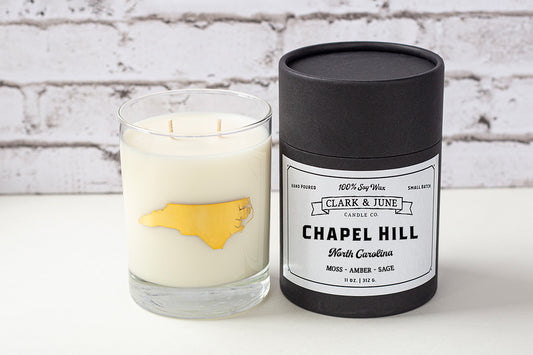 Double wicked soy candle in a 13.5 oz tumbler with the state of North Carolina printed in 22k gold foil on the face. Black cylinder packaging with “Chapel Hill” on the label. SEO Text – Drinking glass, soy wax candle, North Carolina candle, hand poured, small batch, scented candle, Woman Owned, local candle, Housewarming present, gives back, charity, community candle, becomes a cocktail glass, closing gift.