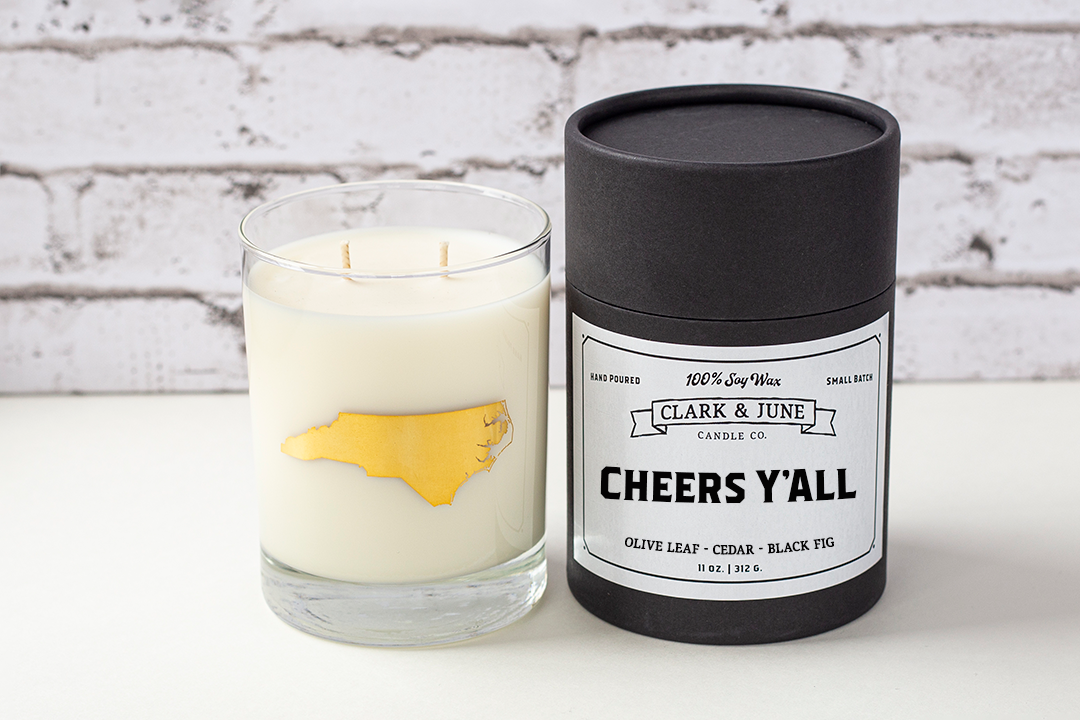 Double wicked soy candle in a 13.5 oz tumbler with the state of North Carolina printed in 22k gold foil on the face. Black cylinder packaging with “Cheers Y’all” on the label. SEO Text – Drinking glass, soy wax candle, North Carolina candle, hand poured, small batch, scented candle, Woman Owned, local candle, Housewarming present, gives back, charity, community candle, becomes a cocktail glass, closing gift.