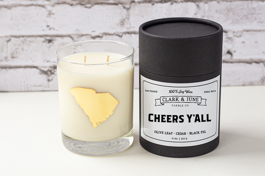 Double wicked soy candle in a 13.5 oz tumbler with the state of South Carolina printed in 22k gold foil on the face. Black cylinder packaging with “Cheers Y’All” on the label. SEO Text – Drinking glass, soy wax candle, South Carolina candle, hand poured, small batch, scented candle, Woman Owned, local candle, Housewarming present, gives back, charity, community candle, becomes a cocktail glass, closing gift.
