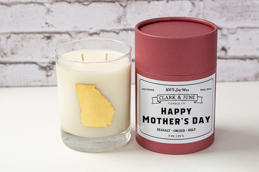 Double wicked soy candle in a 13.5 oz tumbler with the state of Georgia printed in 22k gold foil on the face. Black cylinder packaging with “Happy Mother’s Day” on the label. SEO Text – Drinking glass, soy wax candle, Georgia candle, hand poured, small batch, scented candle, Woman Owned, local candle, Housewarming present, gives back, charity, community candle, becomes a cocktail glass, closing gift. 