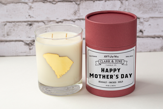 Double wicked soy candle in a 13.5 oz tumbler with the state of South Carolina printed in 22k gold foil on the face. Black cylinder packaging with “Happy Mother’s Day” on the label. SEO Text – Drinking glass, soy wax candle, South Carolina candle, hand poured, small batch, scented candle, Woman Owned, local candle, Housewarming present, gives back, charity, community candle, becomes a cocktail glass, closing gift.