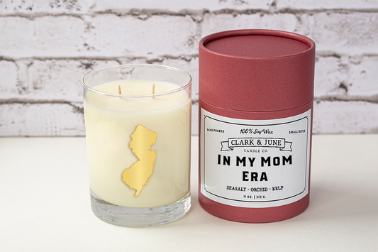 Double wicked soy candle in a 13.5 oz tumbler with the state of New Jersey printed in 22k gold foil on the face. Black cylinder packaging with “In My Mom Era” on the label. SEO Text – Drinking glass, soy wax candle, New Jersey candle, hand poured, small batch, scented candle, Woman Owned, local candle, Housewarming present, gives back, charity, community candle, becomes a cocktail glass, closing gift. Mother's Day Candle