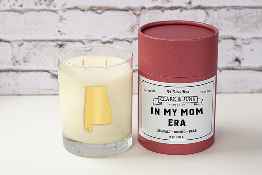 Double wicked soy candle in a 13.5 oz tumbler with the state of Alabama printed in 22k gold foil on the face. Black cylinder packaging with “In My Mom Era” on the label. SEO Text – Drinking glass, soy wax candle, Alabama candle, hand poured, small batch, scented candle, Woman Owned, local candle, Housewarming present, gives back, charity, community candle, becomes a cocktail glass, closing gift. Mother's Day Candle