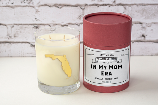 Double wicked soy candle in a 13.5 oz tumbler with the state of Florida printed in 22k gold foil on the face. Black cylinder packaging with “In My Mom Era” on the label. SEO Text – Drinking glass, soy wax candle, Florida candle, hand poured, small batch, scented candle, Woman Owned, local candle, Housewarming present, gives back, charity, community candle, becomes a cocktail glass, closing gift. Mother's Day Candle
