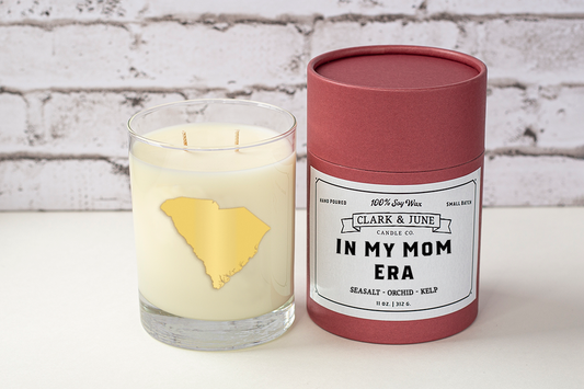 Double wicked soy candle in a 13.5 oz tumbler with the state of South Carolina printed in 22k gold foil on the face. Black cylinder packaging with “In My Mom Era” on the label. SEO Text – Drinking glass, soy wax candle, South Carolina candle, hand poured, small batch, scented candle, Woman Owned, local candle, Housewarming present, gives back, charity, community candle, becomes a cocktail glass, closing gift. Mother's Day Candle 