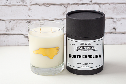 Double wicked soy candle in a 13.5 oz tumbler with the state of North Carolina printed in 22k gold foil on the face. Black cylinder packaging with “North Carolina” on the label. SEO Text – Drinking glass, soy wax candle, North Carolina candle, hand poured, small batch, scented candle, Woman Owned, local candle, Housewarming present, gives back, charity, community candle, becomes a cocktail glass, closing gift.