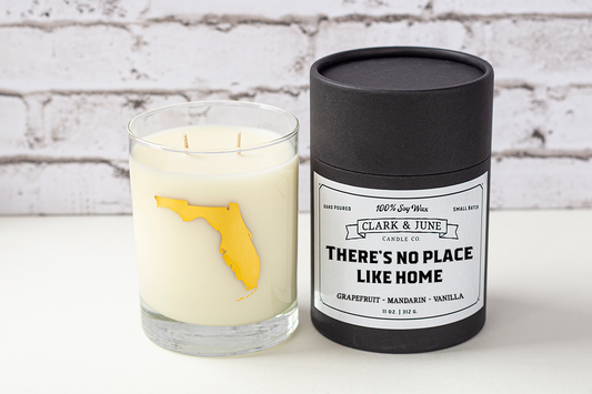 Double wicked soy candle in a 13.5 oz tumbler with the state of Florida printed in 22k gold foil on the face. Black cylinder packaging with "There's No Place Like Home" on the label. SEO Text – Drinking glass, soy wax candle, Florida candle, hand poured, small batch, scented candle, Woman Owned, local candle, Housewarming present, gives back, charity, community candle, becomes a cocktail glass, closing gift. 