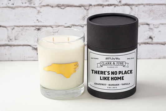 Double wicked soy candle in a 13.5 oz tumbler with the state of North Carolina printed in 22k gold foil on the face. Black cylinder packaging with "There's No Place Like Home"on the label. SEO Text – Drinking glass, soy wax candle, North Carolina candle, hand poured, small batch, scented candle, Woman Owned, local candle, Housewarming present, gives back, charity, community candle, becomes a cocktail glass, closing gift.